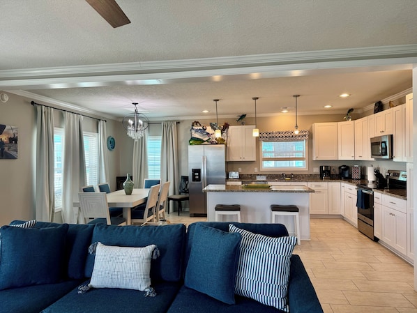 Una sala de estar moderna con un sofá azul oscuro, un comedor con una mesa blanca y una cocina con armarios blancos y electrodomésticos de acero inoxidable.