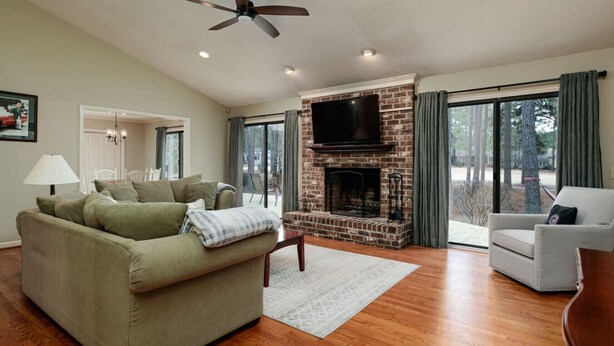 A living room with a brick fireplace, a ceiling fan, and a large window with curtains. - Pinehurst No. 6 Golf Front Getaway (Pinehurst)