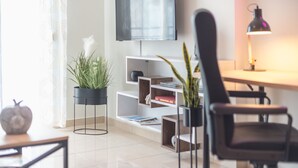 A modern living room with a flat-screen TV, a desk with a lamp, a chair, and a bookshelf.