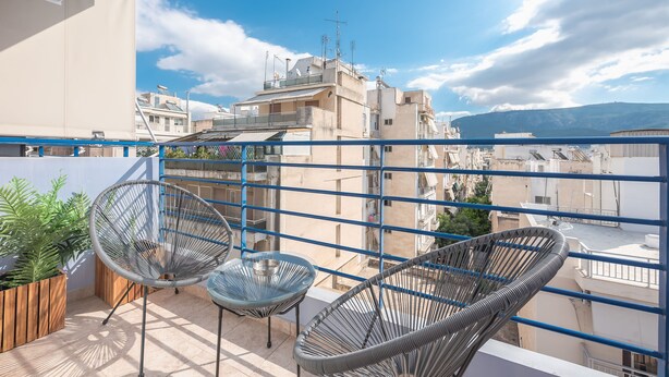 A balcony with two wicker chairs, a wooden planter with green plants, and a view of neighboring buildings and mountains.