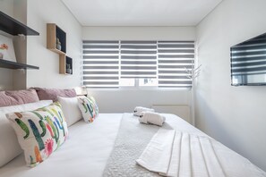 A modern hotel room with a large bed, white bedding, a window with blinds, a mounted TV, and a small shelf with decorative items. - Elegant Home with Balcony by CloudKeys (Athens)