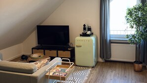 A cozy living room with a flat-screen TV, a vintage-style mini-fridge, a comfortable sofa, a small cart with magazines, and a large window with curtains. - Sweet Spot | Studio apartment close to the city center (Trierweiler)