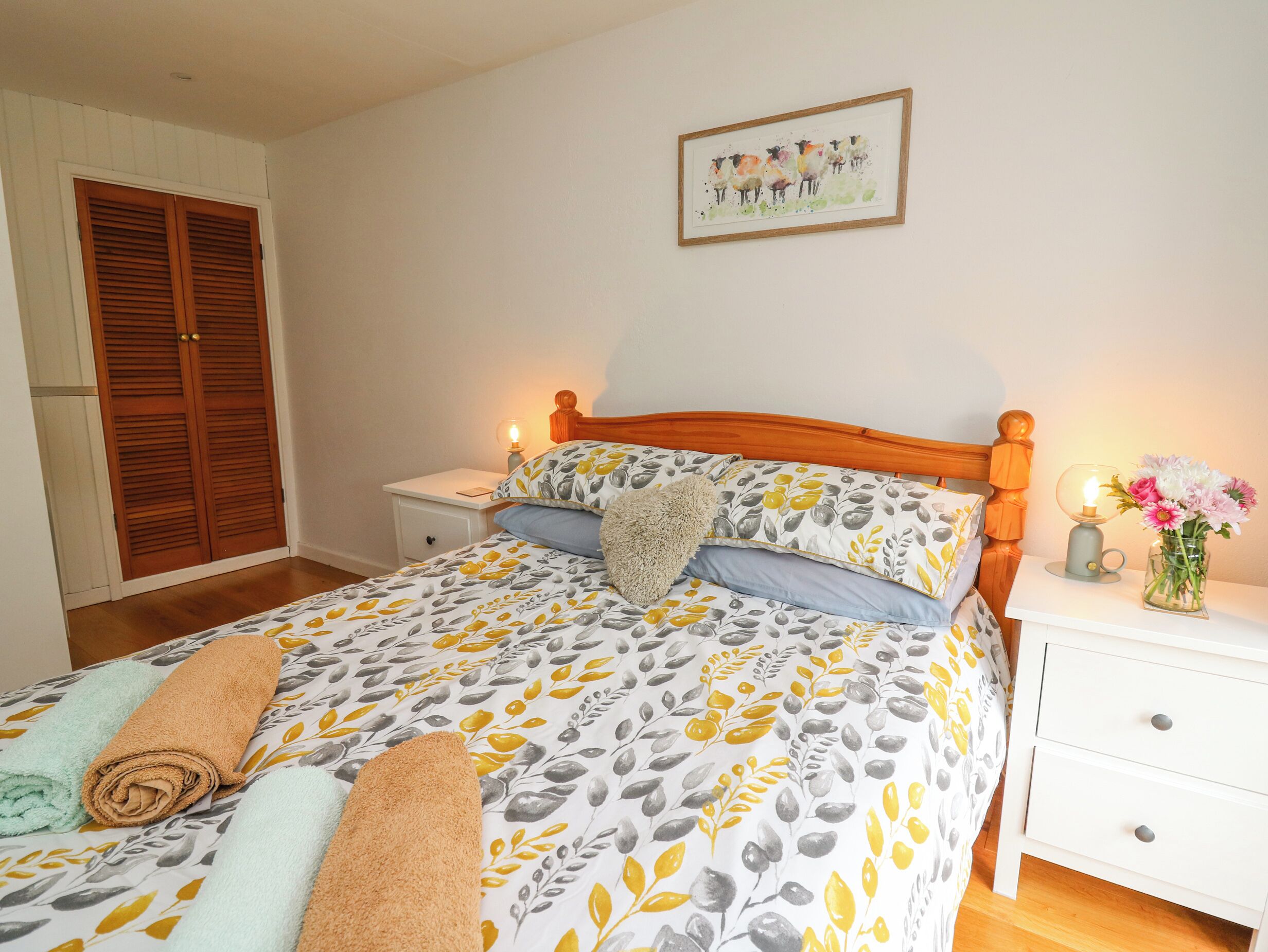 Un dormitorio con cama, mesitas de noche, un armario de madera, una obra de arte enmarco y un jarrón con flores.
