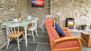 A stone-walled room with a dining table set for two, a wooden bench with cushions, a fireplace, and a painting of a sunset. - Fisherman's Cottage (Llandysul)