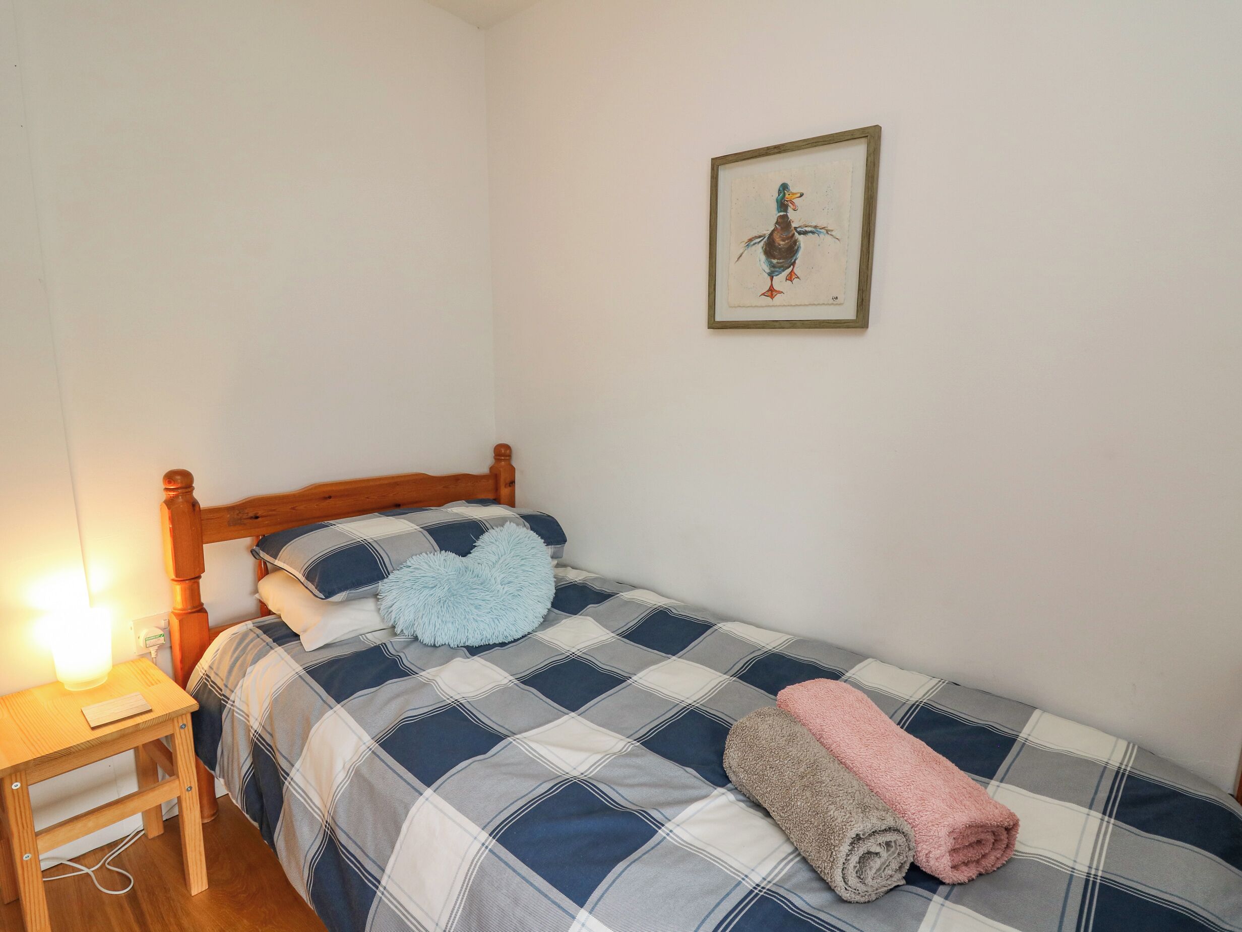A small, neatly made bed with a plaid bedspread and two rolled towels, a wooden nightstand with a lamp, and a framed picture of a duck on the wall.