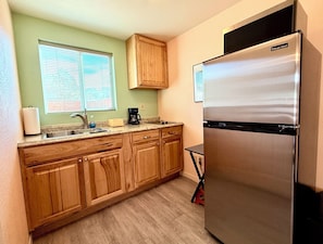 Cocina compacta con armarios de madera, un refrigerador de acero inoxidable, una cafetera y una ventana con persianas.