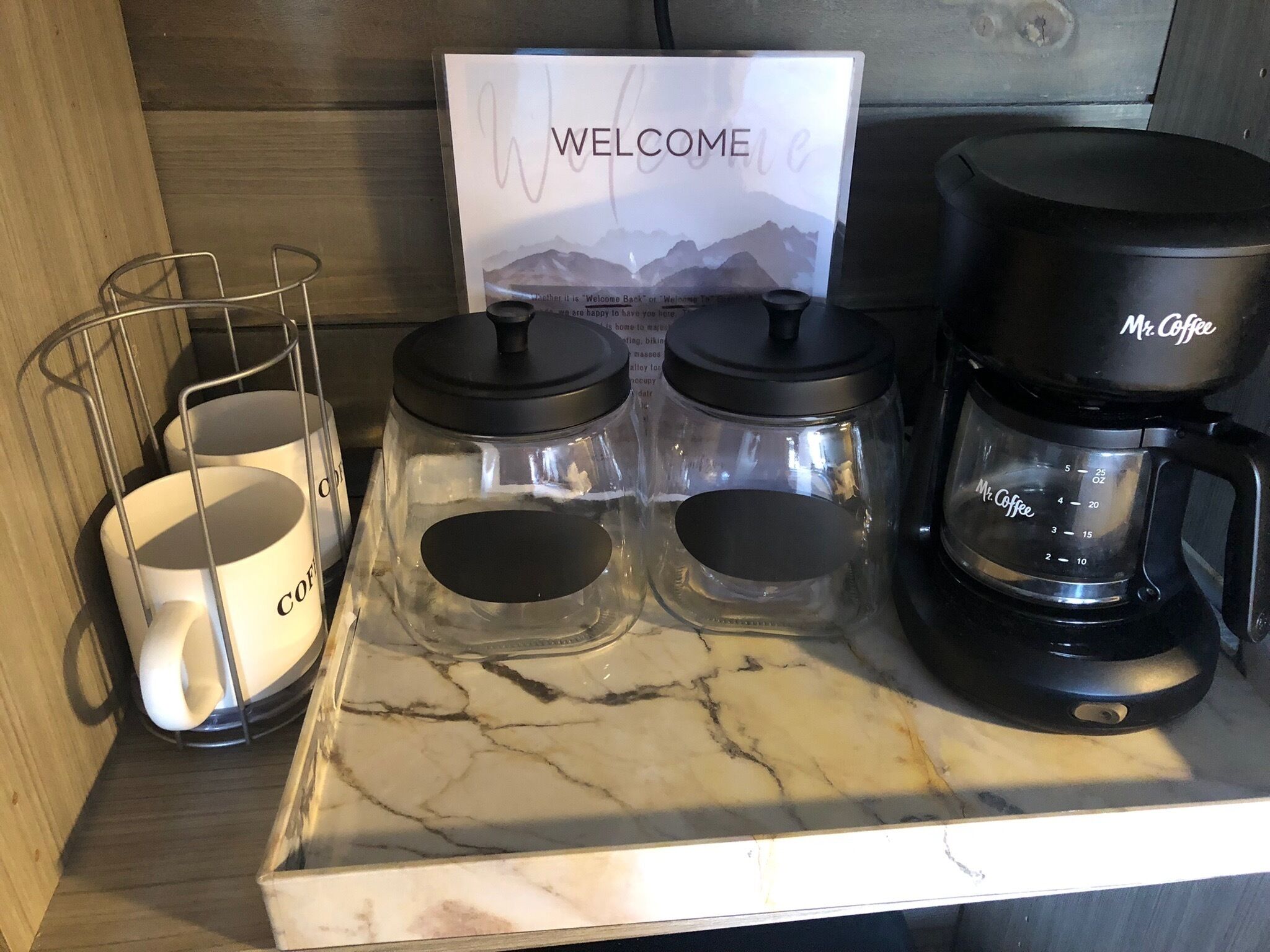 A kitchen counter with a coffee maker, two glass jars with lids, and a welcome sign.