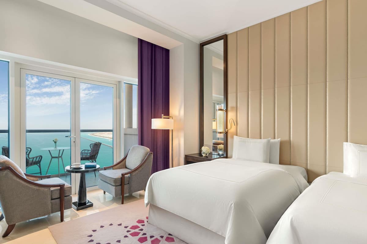 A hotel room with a bed, two chairs, a small table, a lamp, and a large window offering a view of the sea.