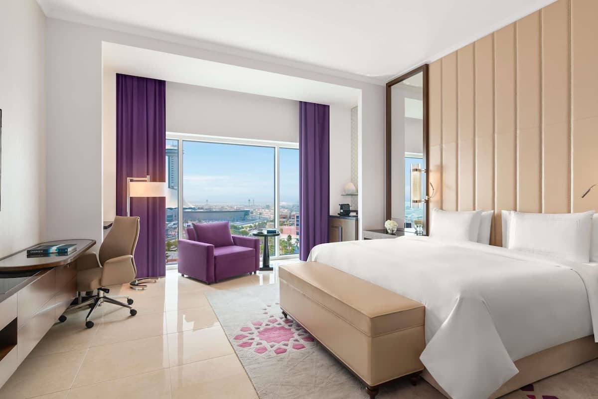 A hotel room with a large bed, a desk, a chair, a sofa, and a view of the city.