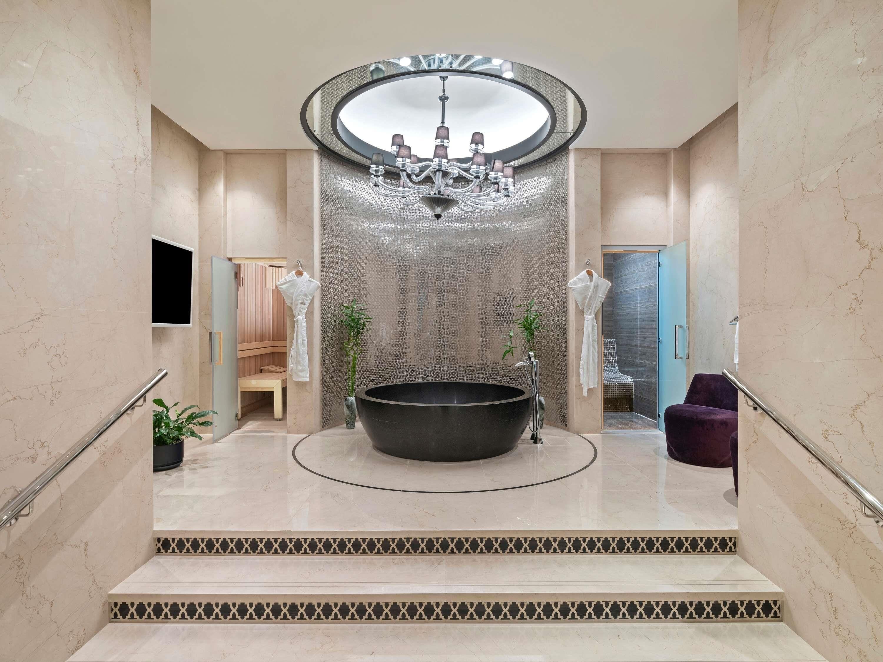 A luxurious bathroom with a large freestanding bathtub, a modern chandelier, and a seating area with a bench and a chair.