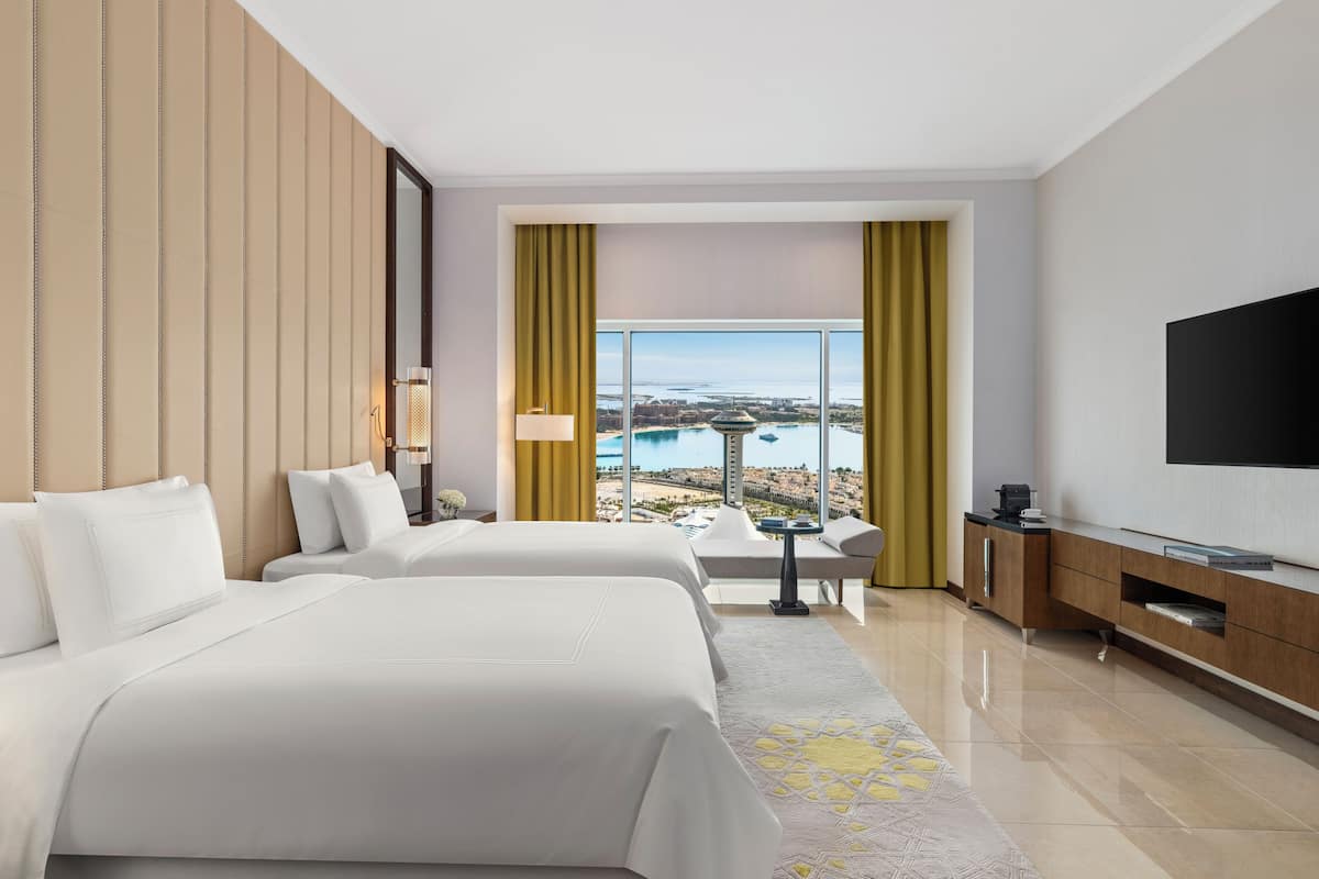 A hotel room with two beds, a TV, a desk, and a view of a cityscape and water.