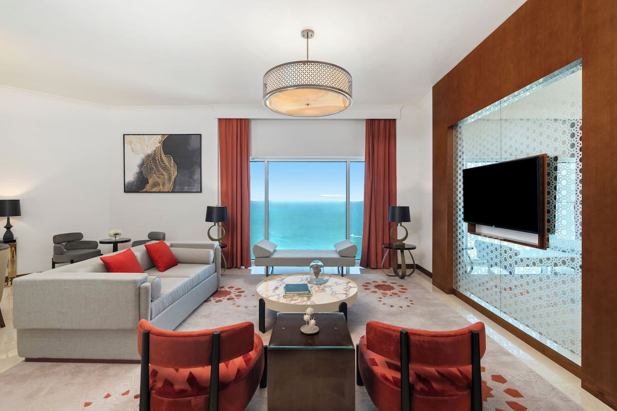 A modern hotel room with a large window offering a view of the sea, a comfortable sofa, a coffee table, and red chairs.