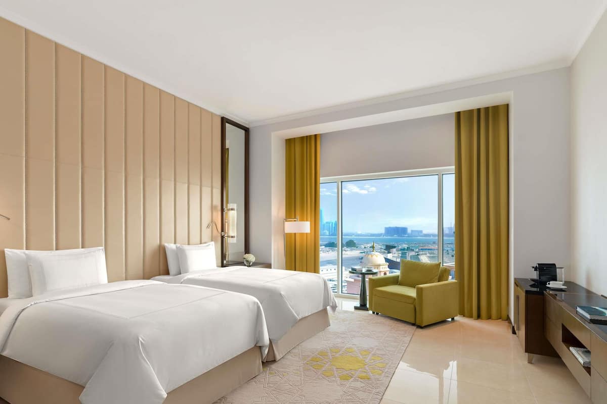 A hotel room with two beds, a sitting area with a chair, a small table, and a view of the city.
