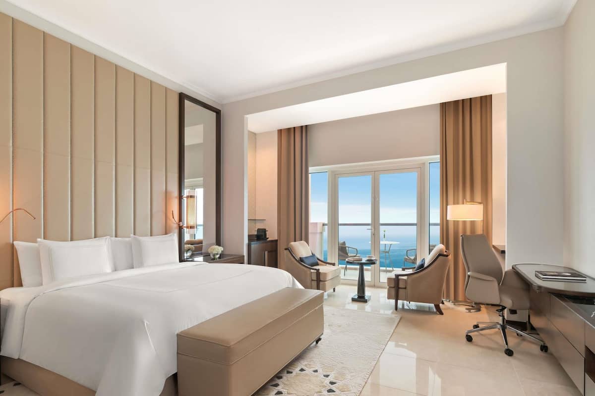 A hotel room with a large bed, a desk, two chairs, a view of the ocean, and a balcony.