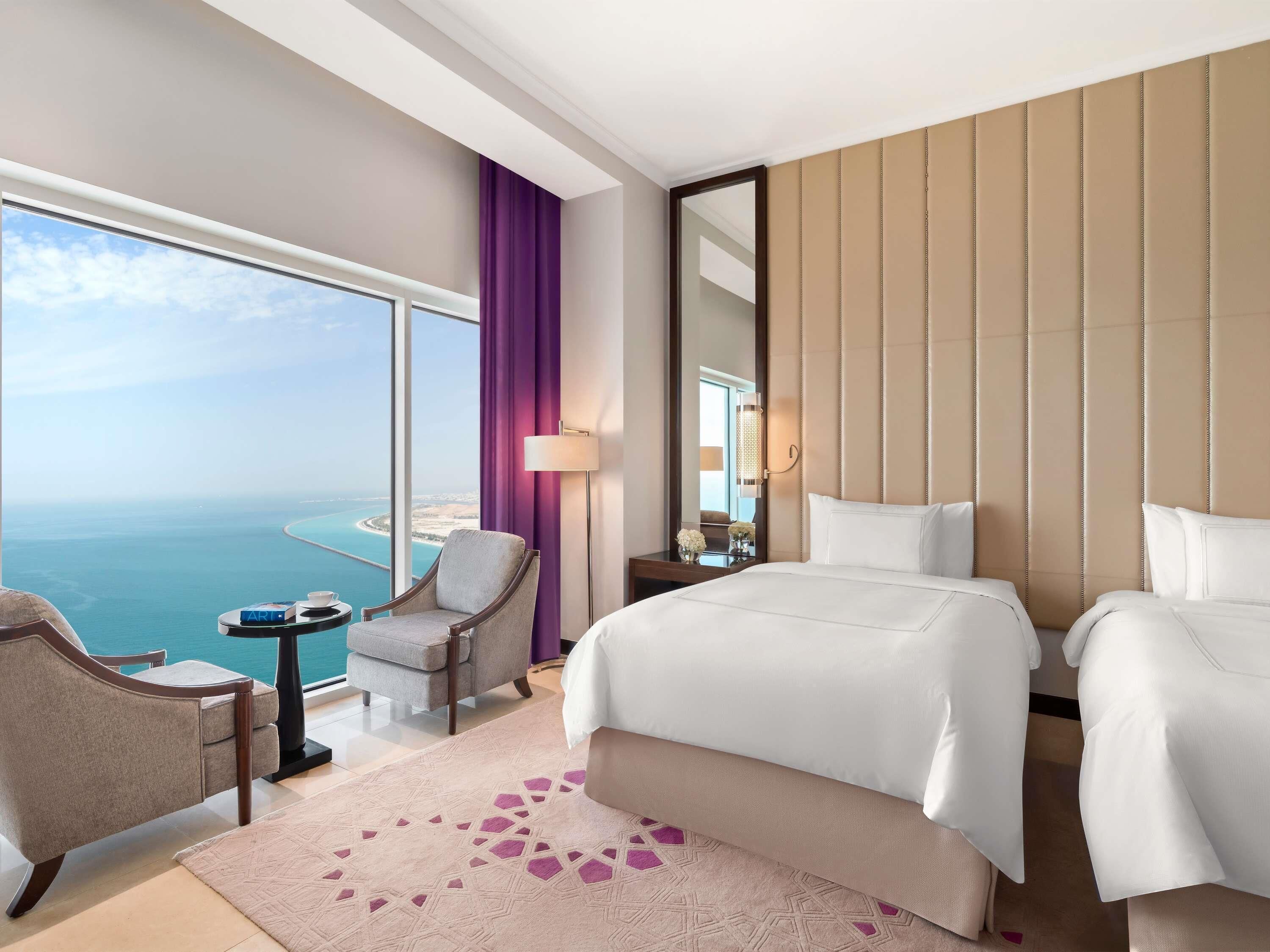 Quarto de hotel com uma cama grande, uma cadeira, uma mesinha, um abajur e vista para o mar.