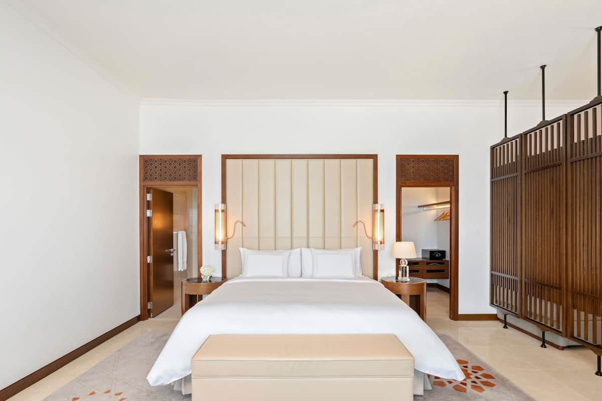 A hotel room with a large bed, bedside tables, a bench, a wardrobe, and a mirror.