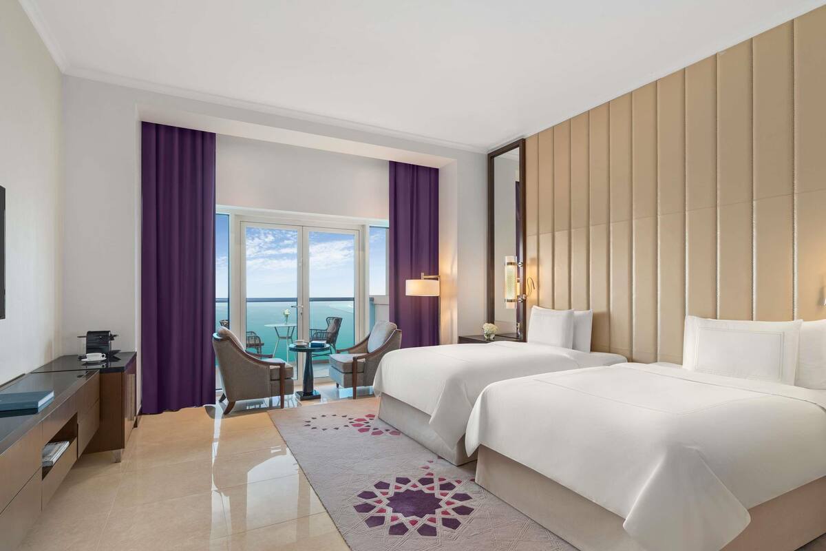 A hotel room with a balcony, two beds, a desk, and a chair.