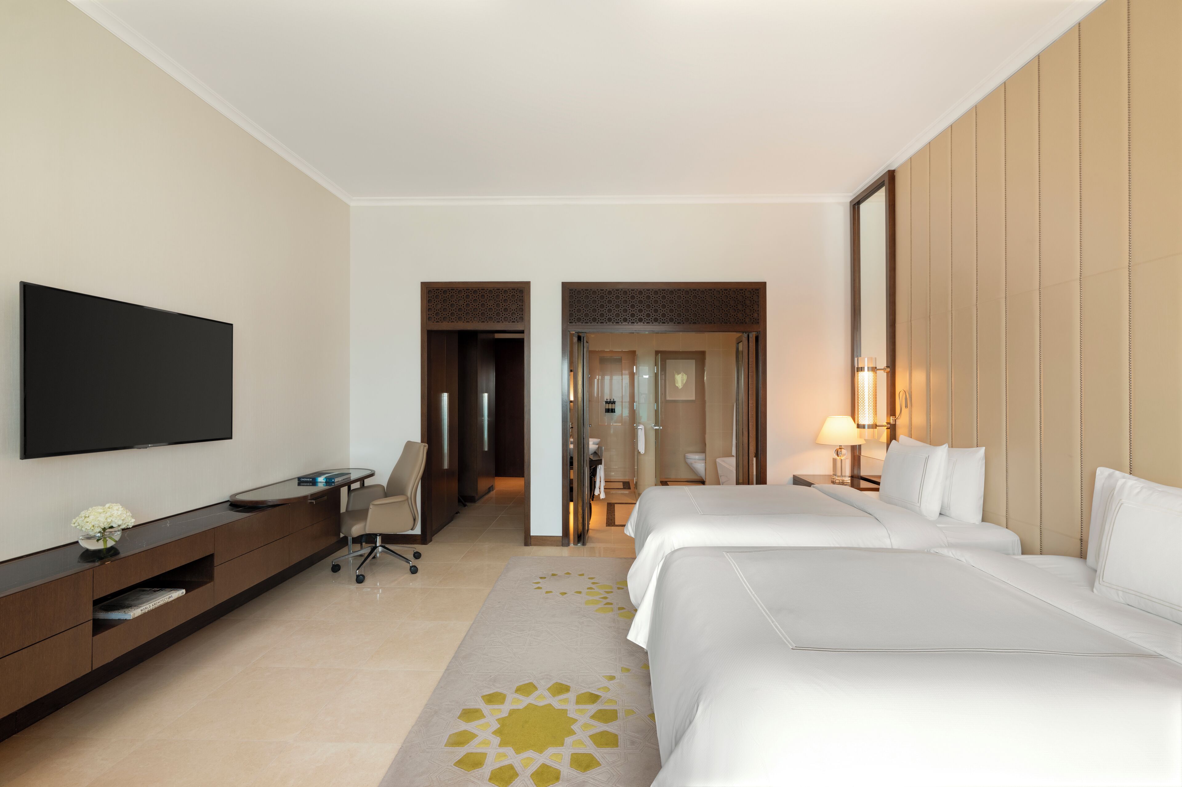 Une chambre d’hôtel équipée d’une télévision à écran plat, d’un bureau avec une chaise, de deux lits, d’une salle de bain et d’un couloir menant à une autre chambre.
