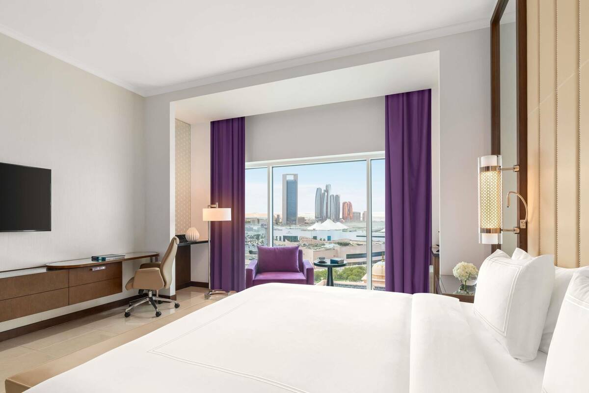 A hotel room with a large bed, a desk, a chair, a TV, and a view of a cityscape.