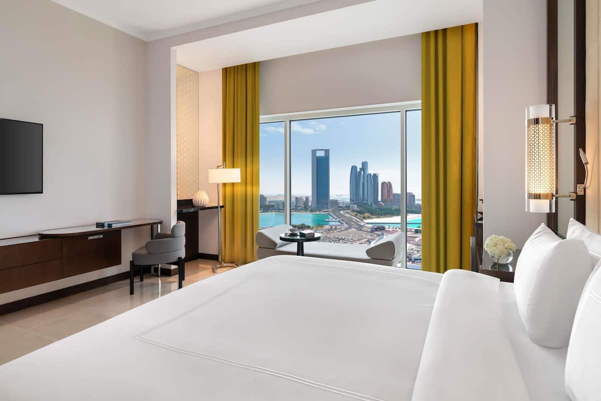 A hotel room with a large bed, a desk, a chair, a TV, a lamp, and a view of a cityscape.