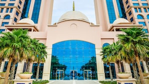 A hotel entrance with a central fountain, tall buildings with blue-tinted windows, and palm trees. - Rixos Marina Abu Dhabi (Abu Dhabi)