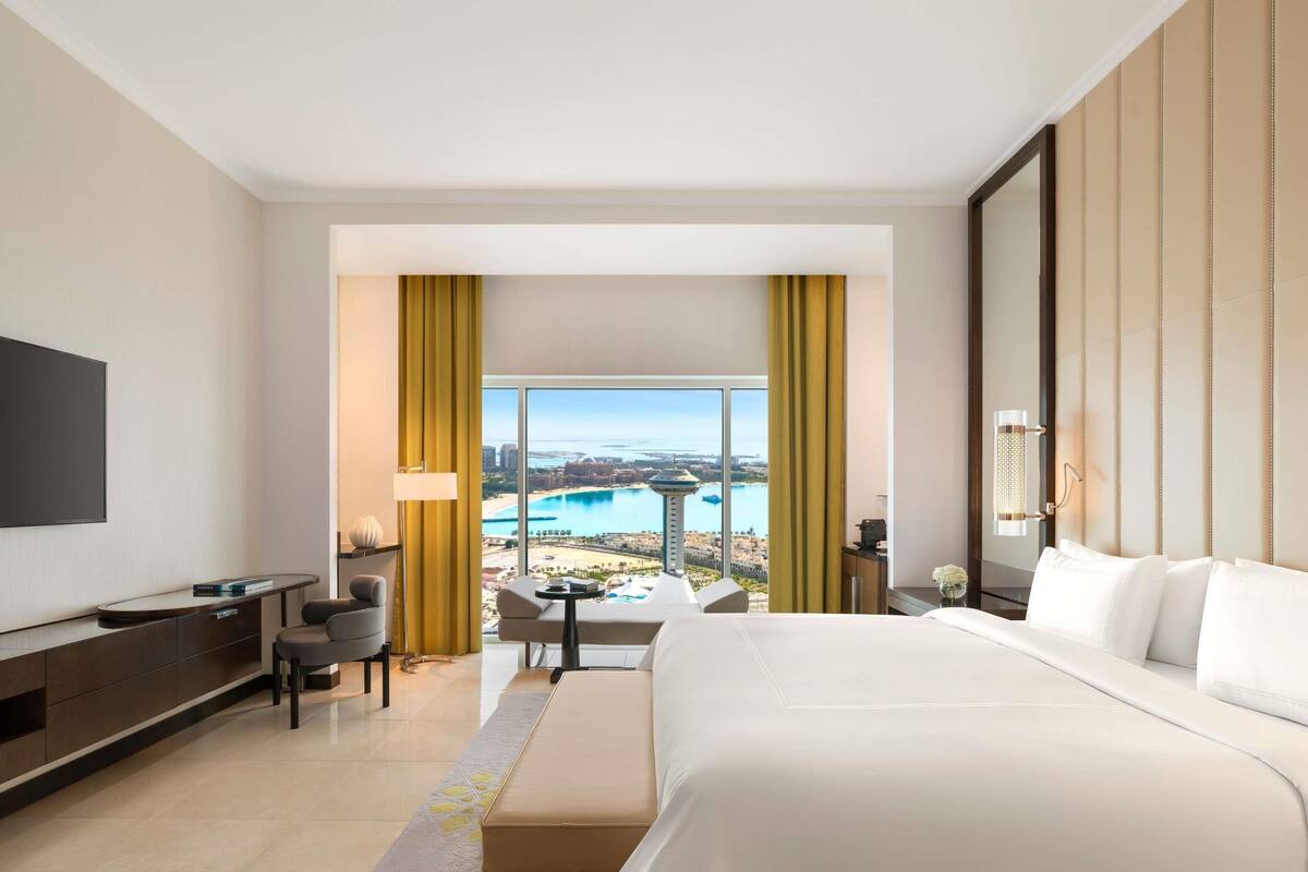 A hotel room with a large bed, a desk, a chair, and a view of a beach and ocean.