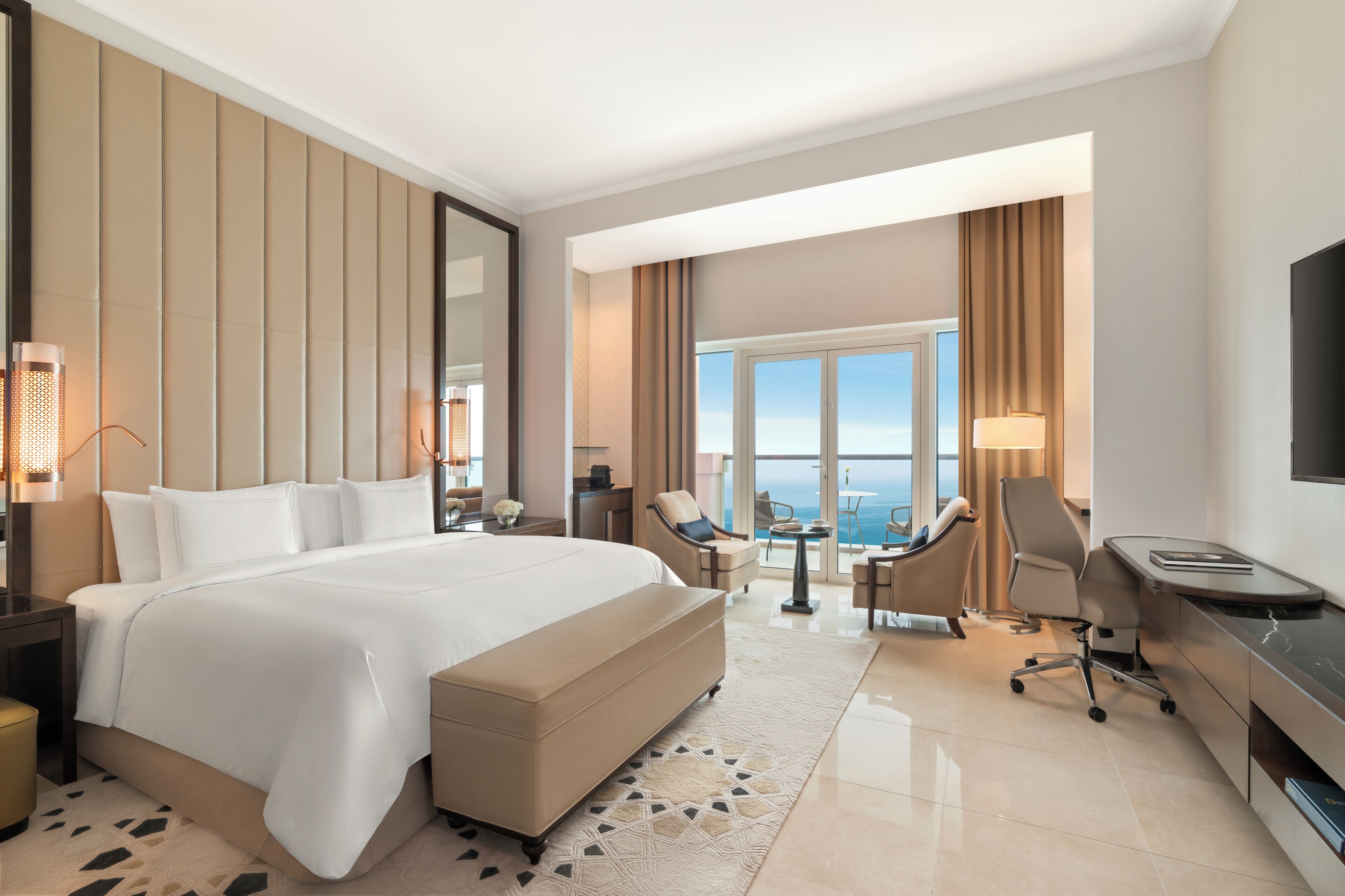 A modern hotel room with a large bed, a desk, a chair, and a view of the ocean.