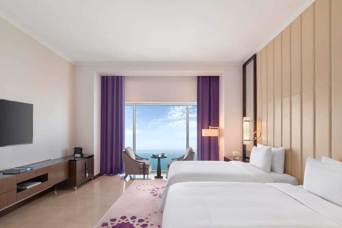 A hotel room with a bed, a desk, a TV, a chair, a table, and a view of the ocean.