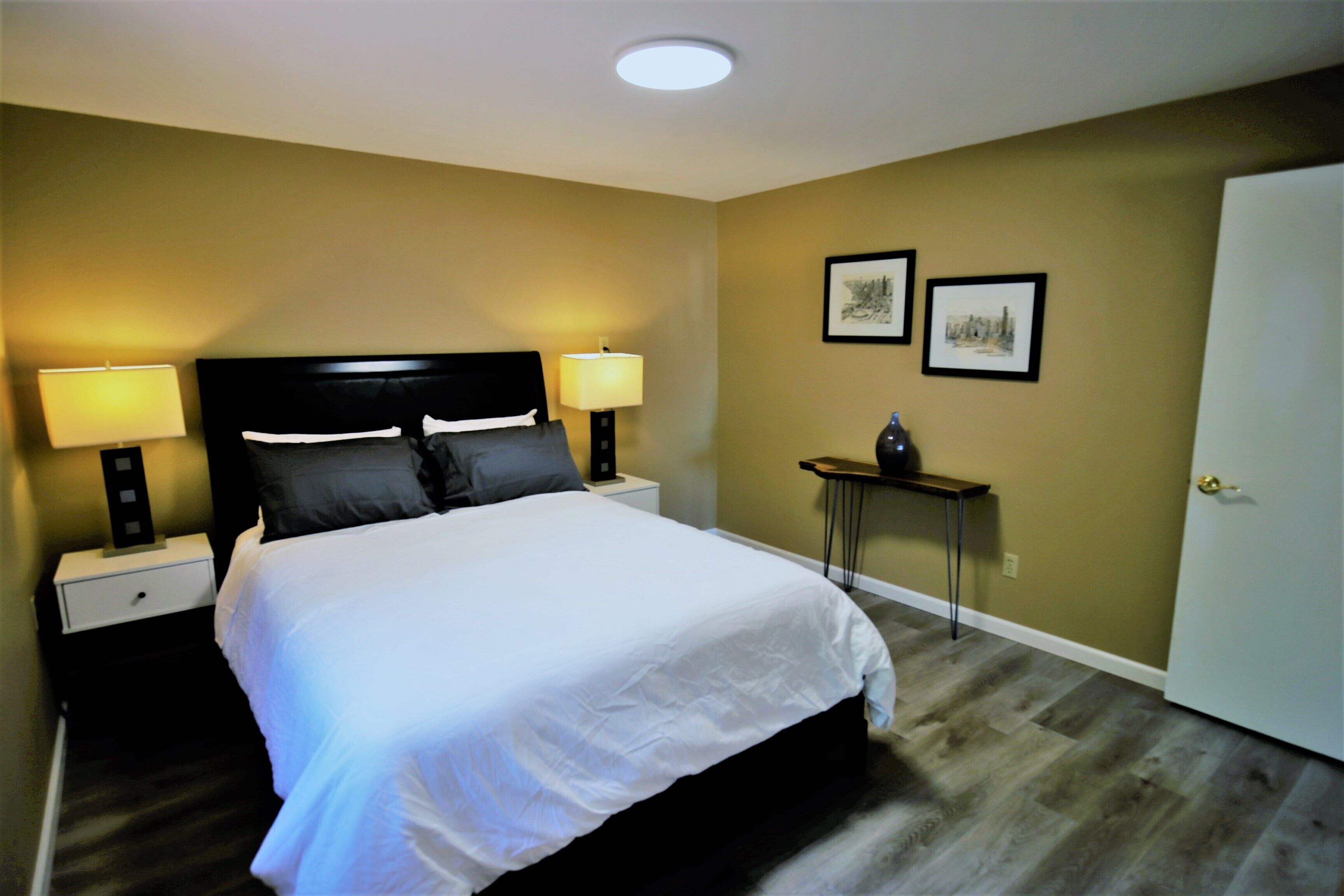 Habitación de hotel con una cama grande, dos mesitas de noche con lámparas, una pequeña mesa con un florero y dos cuadros en la pared.