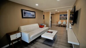 A modern living room with a white sofa, a glass coffee table, and a small wooden side table. There is a framed picture on the wall and a flat-screen TV mounted on the wall.