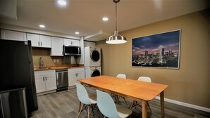 A modern kitchen with white cabinets, a stainless steel refrigerator, a microwave, and a washing machine. There is a wooden dining table with four chairs and a pendant light above it. A cityscape painting is on the wall.