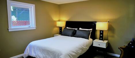 Una cama bien tendida en una habitación con ventana, dos mesitas de noche con lámparas y una silla.