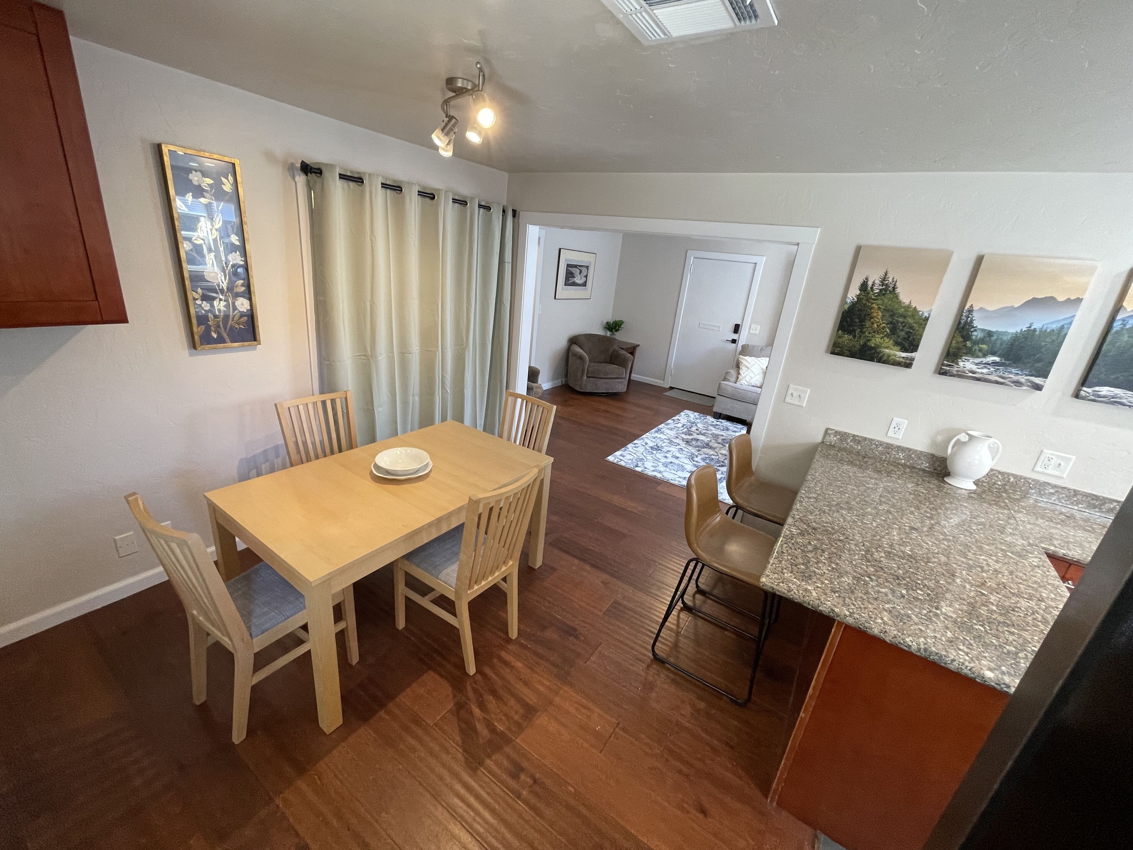 A dining area with a wooden table and chairs, a kitchen with granite countertops, and a living area with a sofa and a plant.