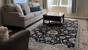 A living room with a beige sofa, a coffee table, a patterned rug, and a view of the outdoors through large windows.