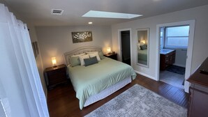 A bedroom with a bed, bedside tables, a nightstand, a window, and a door leading to a bathroom.