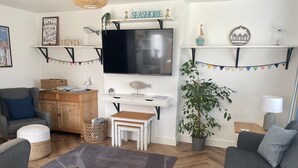 Una sala de estar con televisión, sofás grises, un mueble de madera, un escritorio y una planta en maceta.