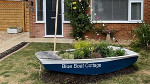 Una casa con techo de tejas, fachada de ladrillo y puerta principal. Frente a la casa, un letrero dice 'Blue Boat Cottage'.
