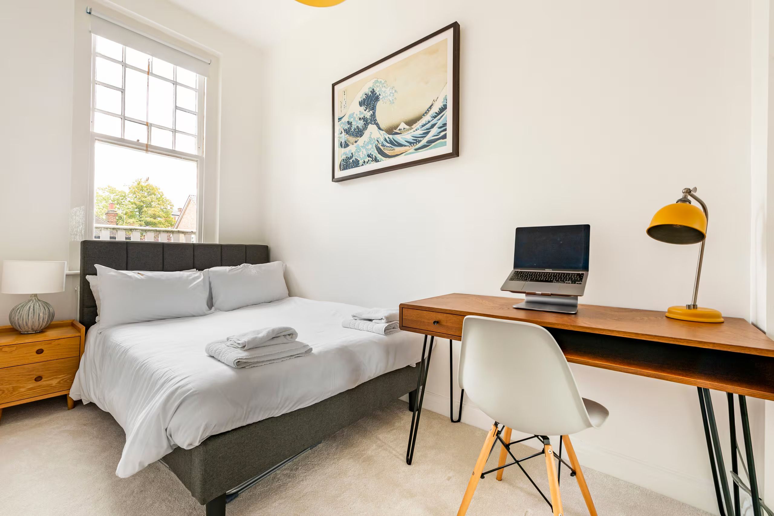 A bedroom with a bed, a desk with a laptop, a chair, a lamp, and a painting on the wall.