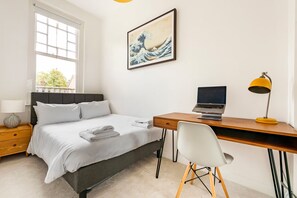 Ein Schlafzimmer mit einem Bett, einem Schreibtisch mit einem Laptop, einem Sessel, einer Lampe und einem Bild an der Wand.