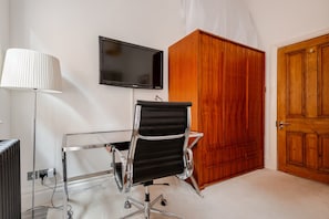 Ein Zimmer mit schwarzem Bürostuhl, höhenverstellbarem Schreibtisch, an der Wand montiertem Fernseher, Holzschrank und einer Tür.