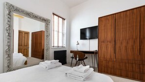 Habitación de hotel con un espejo grande y decorativo, una cama con toallas blancas ordenadas, un armario de madera, un escritorio con una silla y un televisor de pantalla plana montado en la pared.