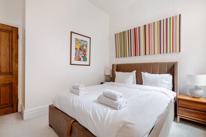 A hotel room with a bed, a nightstand, a lamp, a wooden door, and a colorful abstract painting on the wall. - Luxury Central London 2 x double bed apartment - Hampstead (London)
