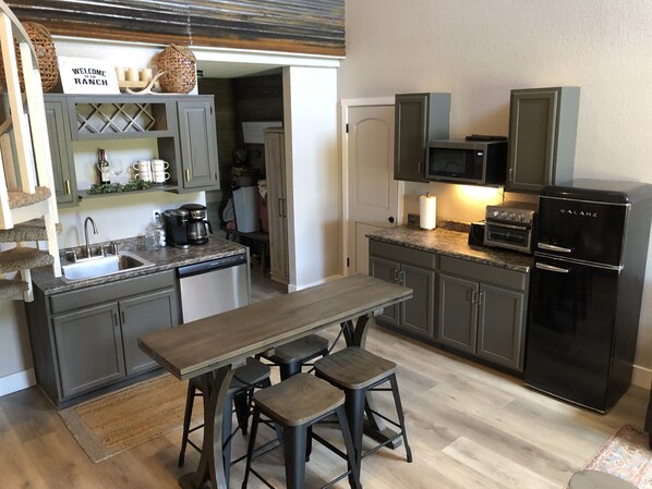 Una cocina con una mesa de madera, taburetes de bar, horno empotrado, microondas y refrigerador.