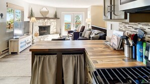 A modern kitchen with a stone fireplace, a dining area with a wooden table, and a view of the outdoors through large windows. - The Tree House - Cozy, mountain cabin (Evergreen)