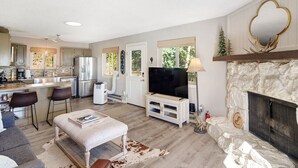 A spacious living area with a fireplace, a television, and a kitchen in the background. - The Tree House - Cozy, mountain cabin (Evergreen)