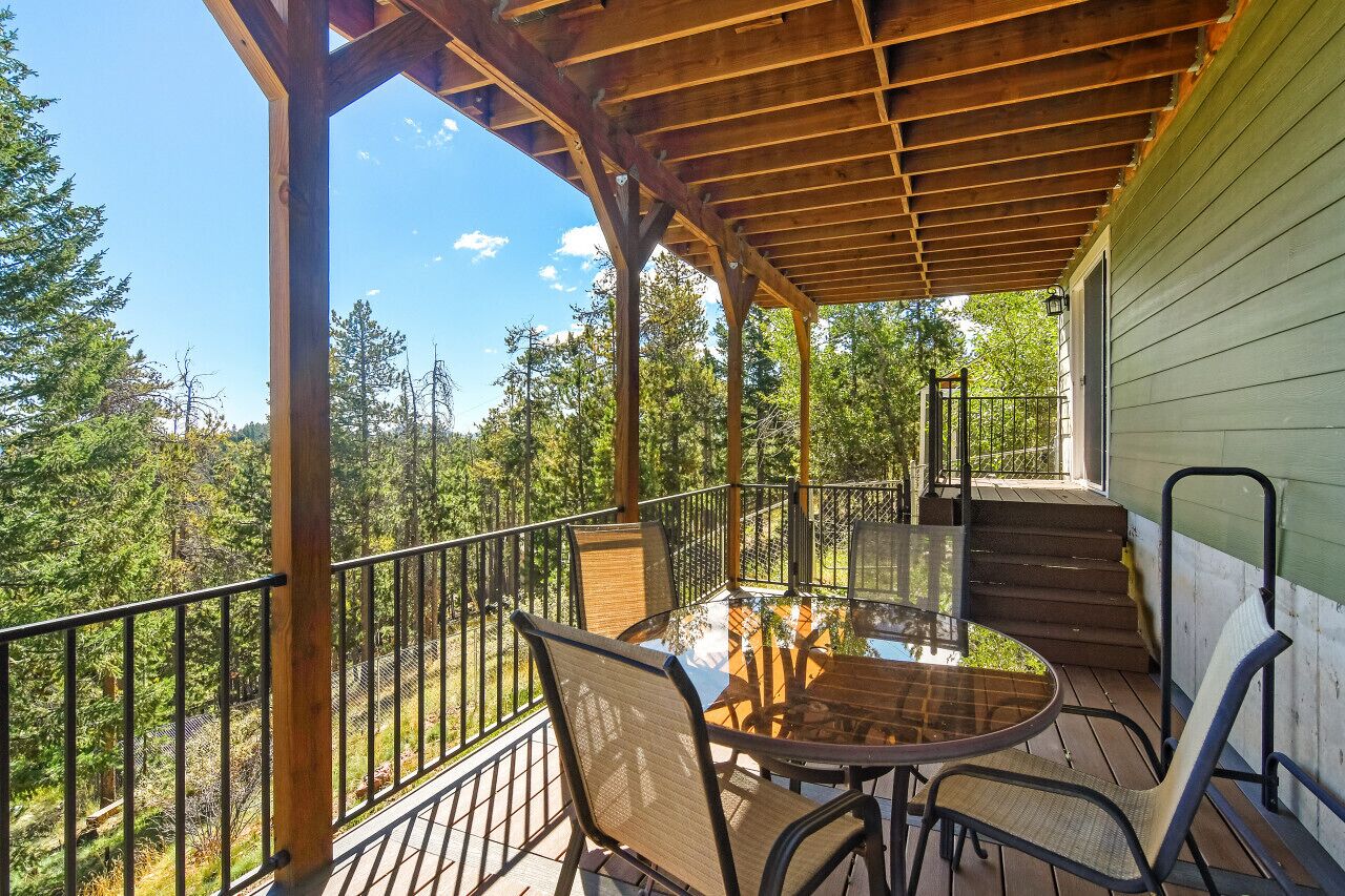 A covered outdoor patio with a round table and chairs, surrounded by a metal railing, overlooking a forested area.