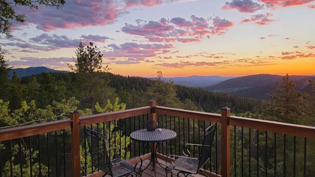 A wooden deck with a table and chairs, overlooking a forested landscape with mountains in the distance. - The Tree House - Cozy, mountain cabin (Evergreen)
