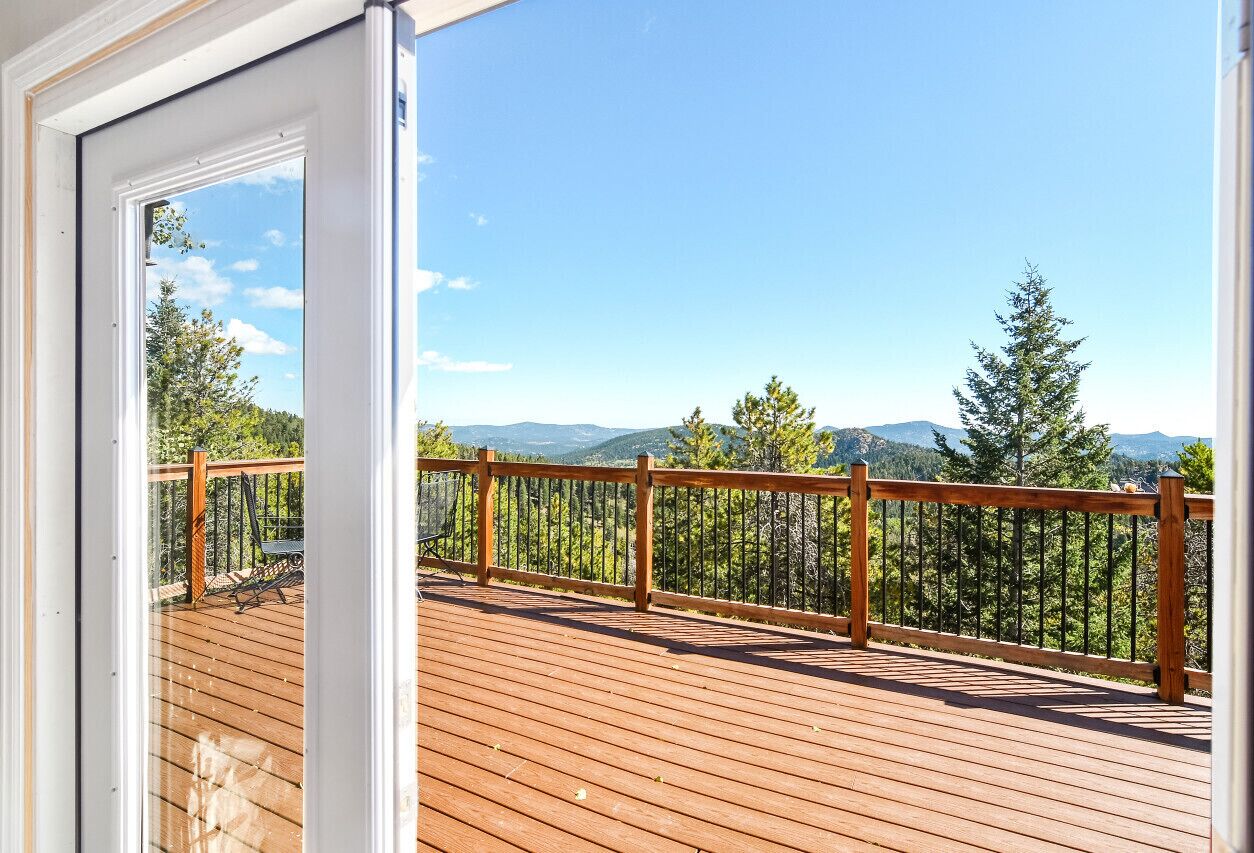 A wooden deck with a railing, overlooking a forested area and distant mountains.