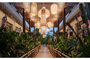 Stroll through a lush garden setting, flanked by tropical plants and trees, with a pathway lined with wooden railings.