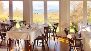 A dining area with tables set for a meal, large windows offering a view of the countryside. - The Colonsay Hotel (Isle of Colonsay)